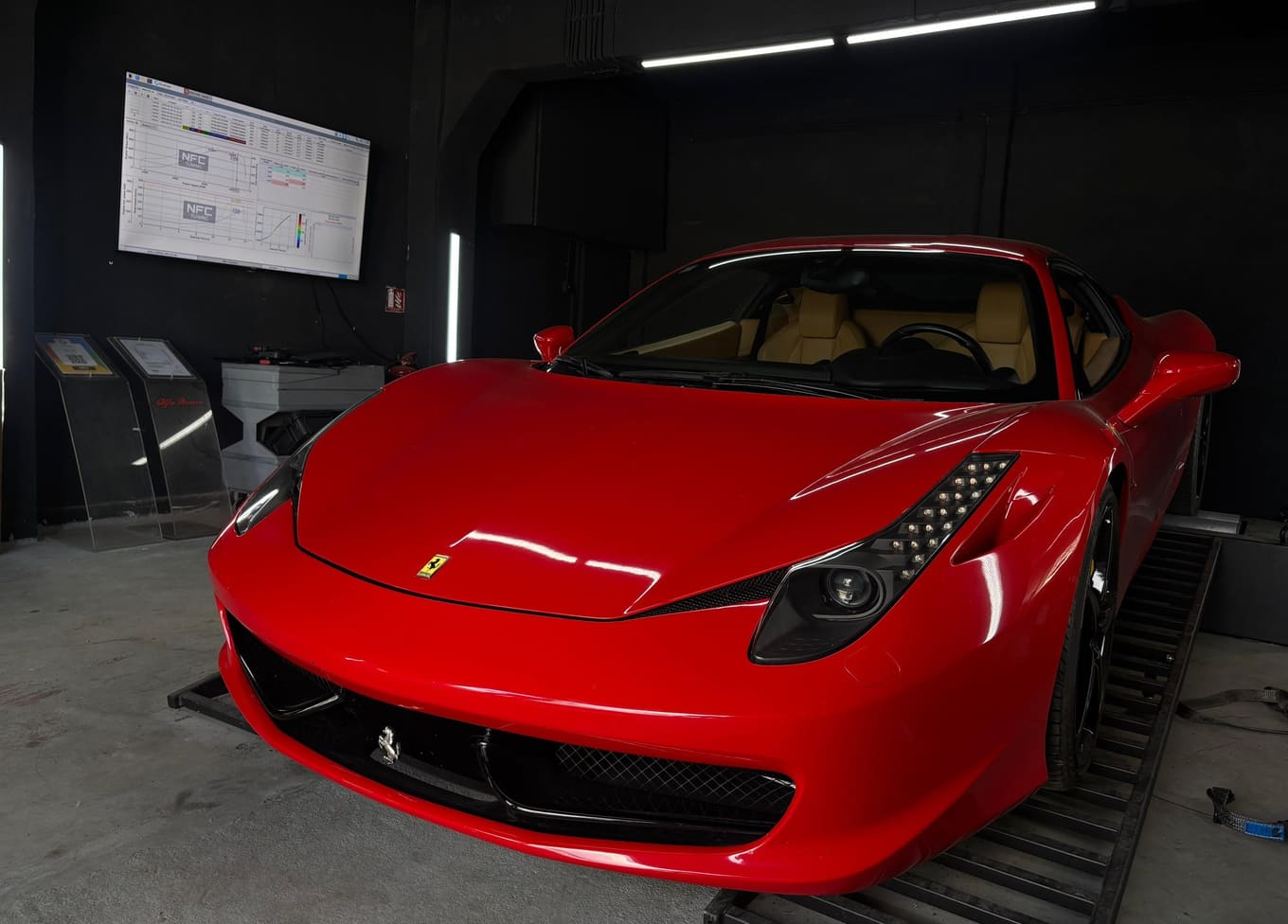 Crveni Ferrari 458 na valjcima u zamračenoj mjernoj komori radionice NFC Tuning, s grafovima snage na zaslonu na zidu